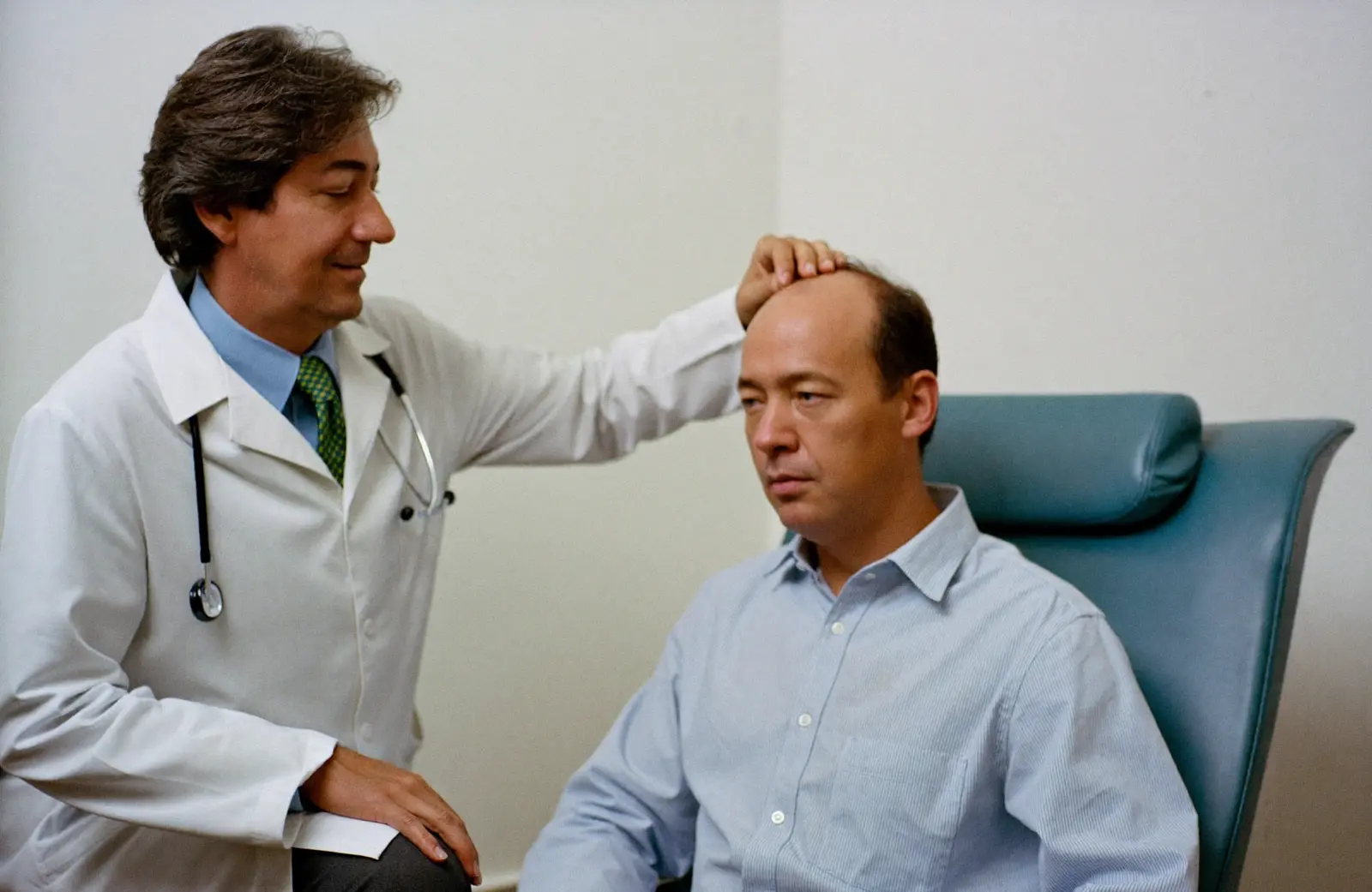 A doctor in a white coat examining the thinning crown of a seated man’s scalp
