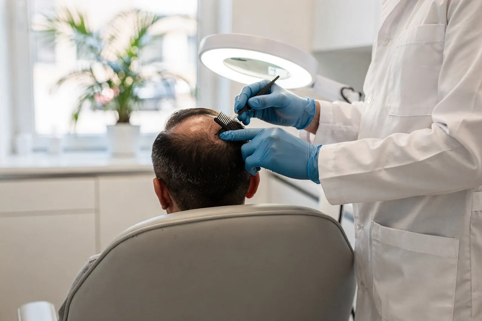 A gloved clinician parts the hair at the thinning crown of a seated patient with a fine comb under a ring magnifier lamp, seen from behind in a bright clinic room