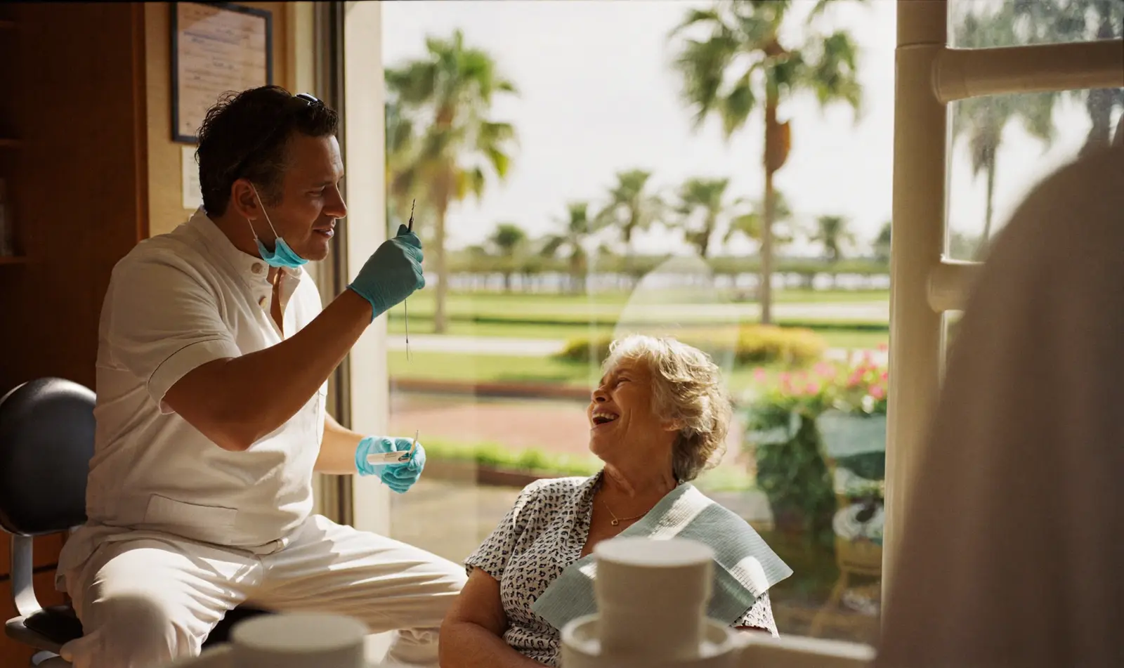 A dentist in white scrubs talking with a smiling older woman in a sunny clinic with palm trees outside