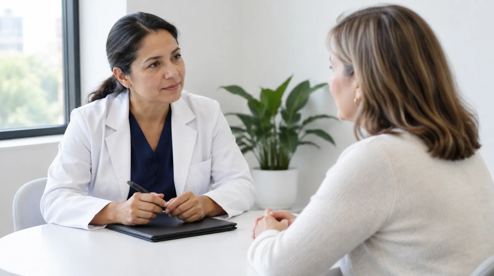 A doctor in a white coat listens to a patient across a small table in a bright clinic office