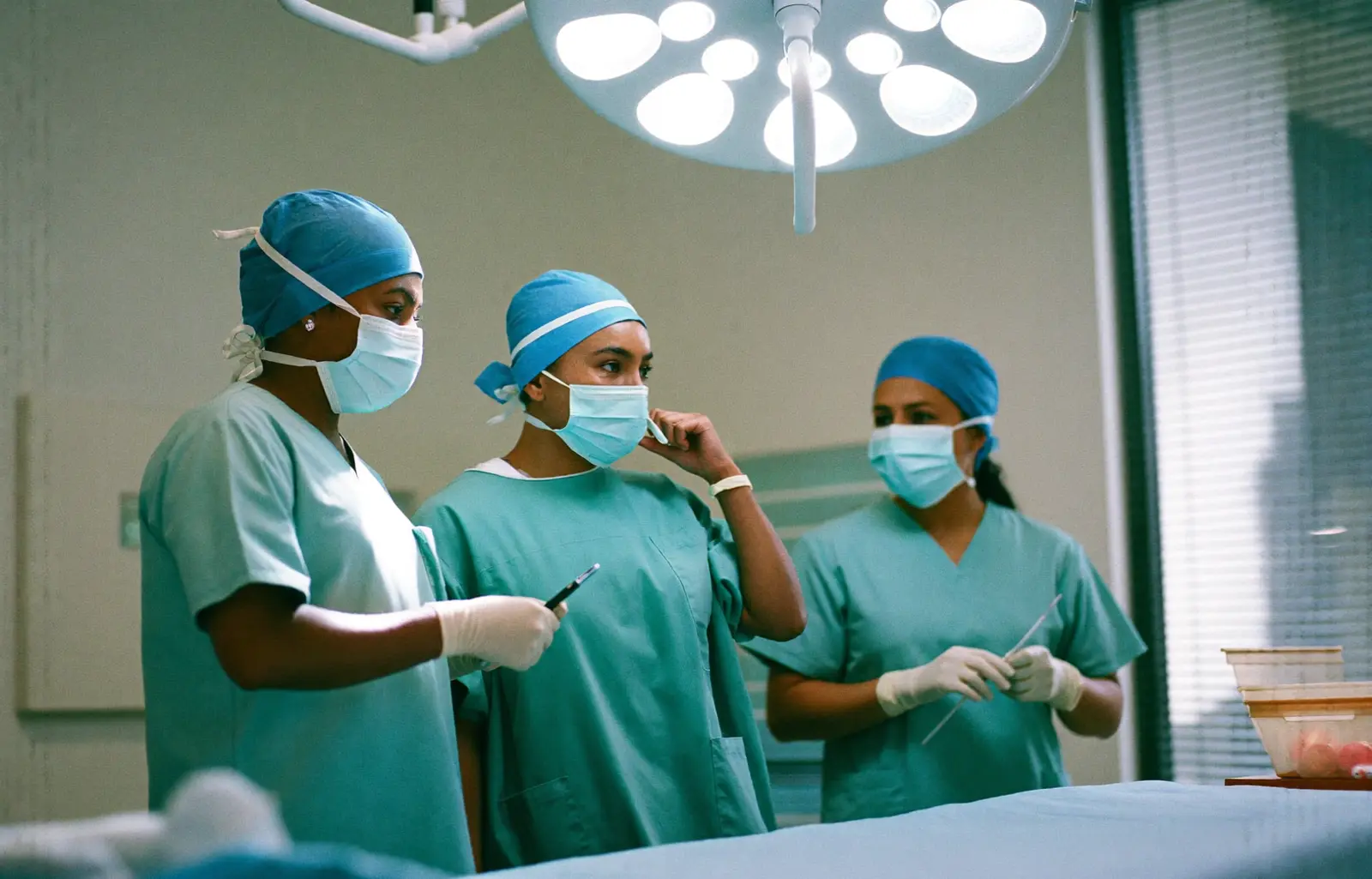 Three members of a surgical team in teal scrubs, caps and masks preparing under the lights of an operating room