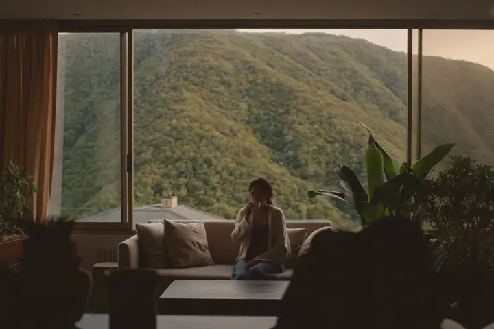 A woman in a cardigan sitting on a sofa by a large window with a green mountainside behind it