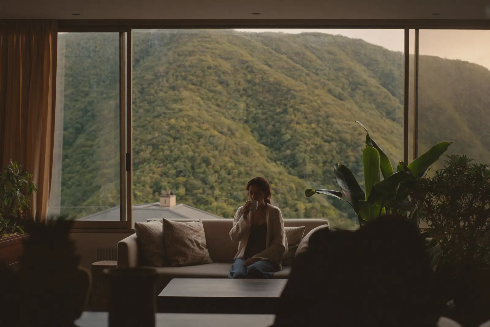 A woman in a cardigan sitting on a sofa by a large window with a green mountainside behind it